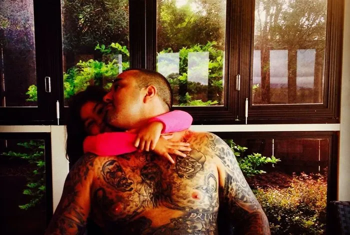 Tattooed parent embraces child in a warm hug by a window with greenery outside.