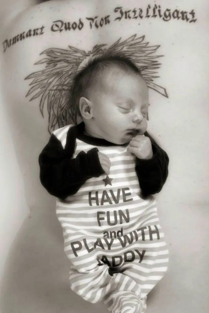 Baby sleeps peacefully on tattooed parent's chest, wearing a striped onesie with a "Have Fun and Play with Daddy" message.