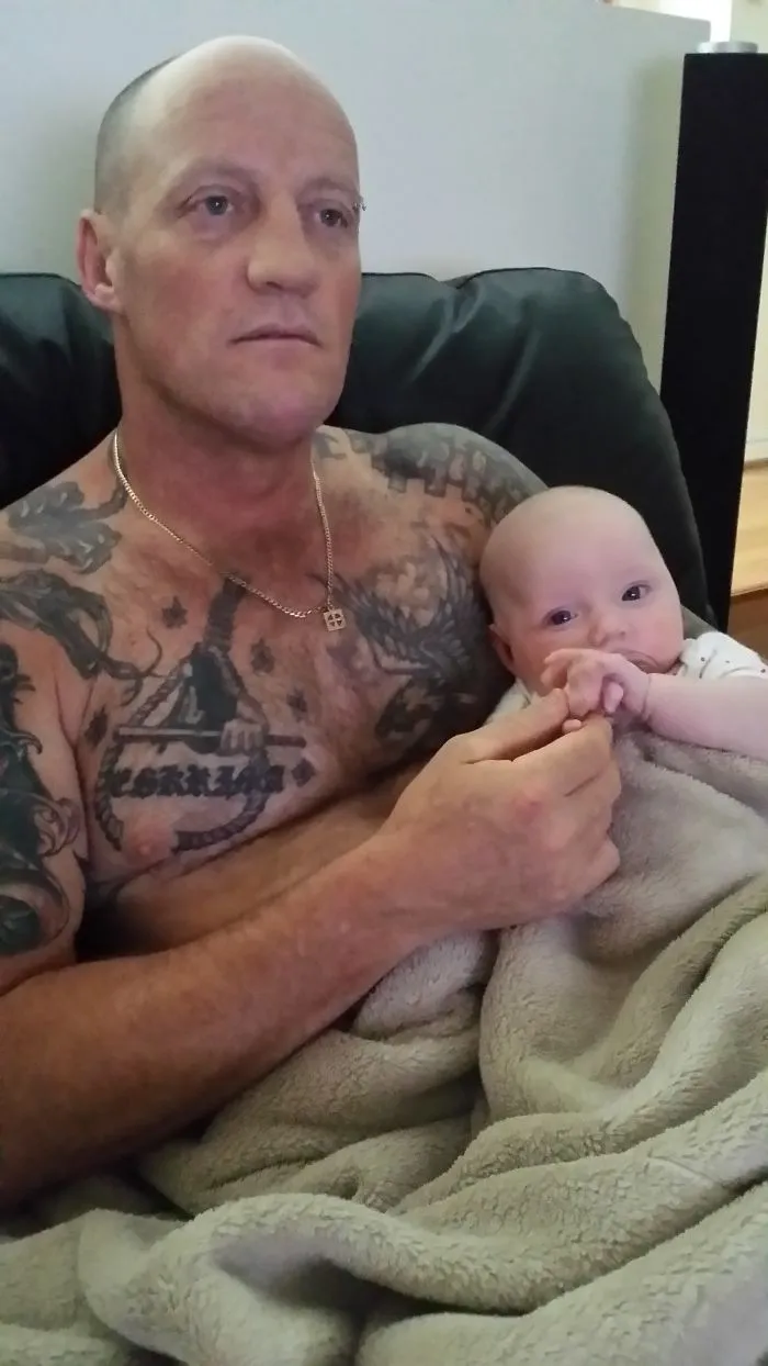 Tattooed parent holding a baby, wrapped in a cozy blanket, seated together on a couch.