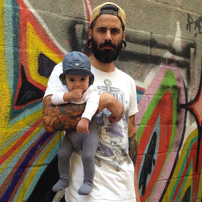 Tattooed parent holds a baby in front of colorful graffiti, both looking at the camera.