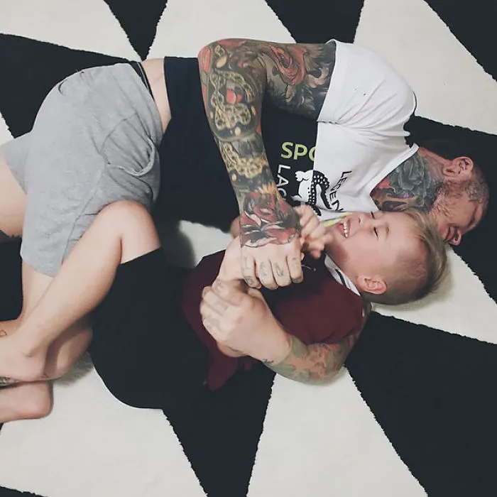 Tattooed parent playing joyfully with a child on a patterned rug, both sharing laughter and closeness.