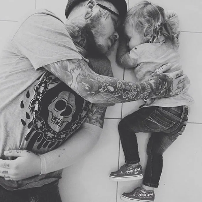 Tattooed parent and child lying together in matching gray shirts, showcasing a tender bond.
