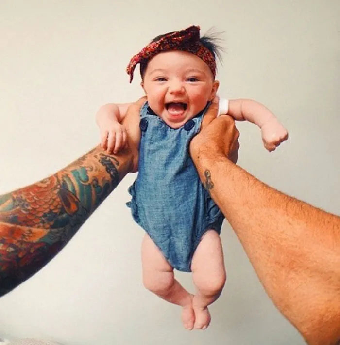 Smiling baby held by tattooed parent, wearing a denim outfit and headband.