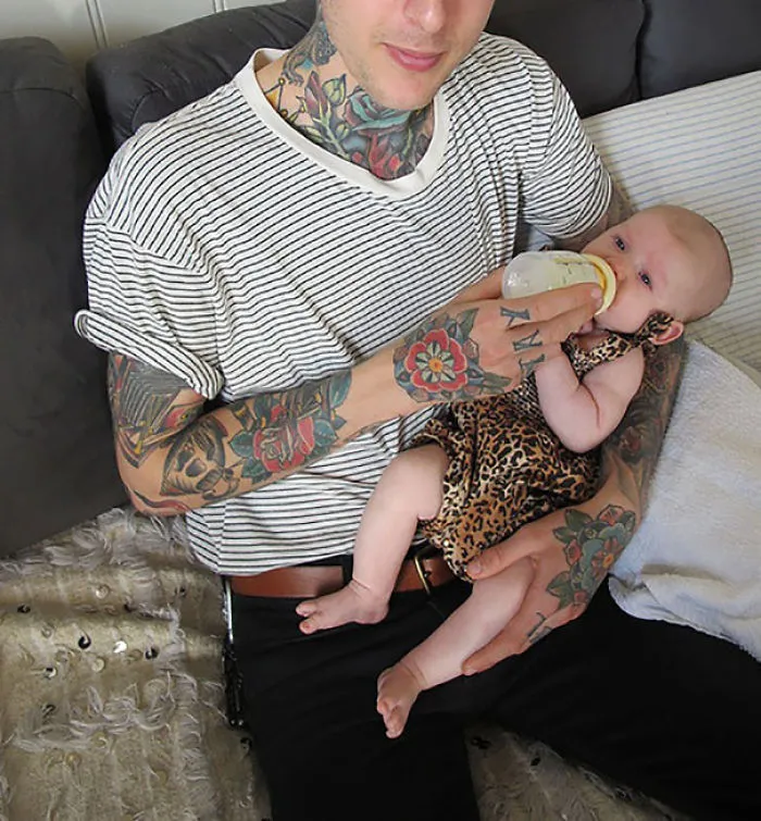 Tattooed parent holding and feeding baby with bottle on a cozy couch setting.