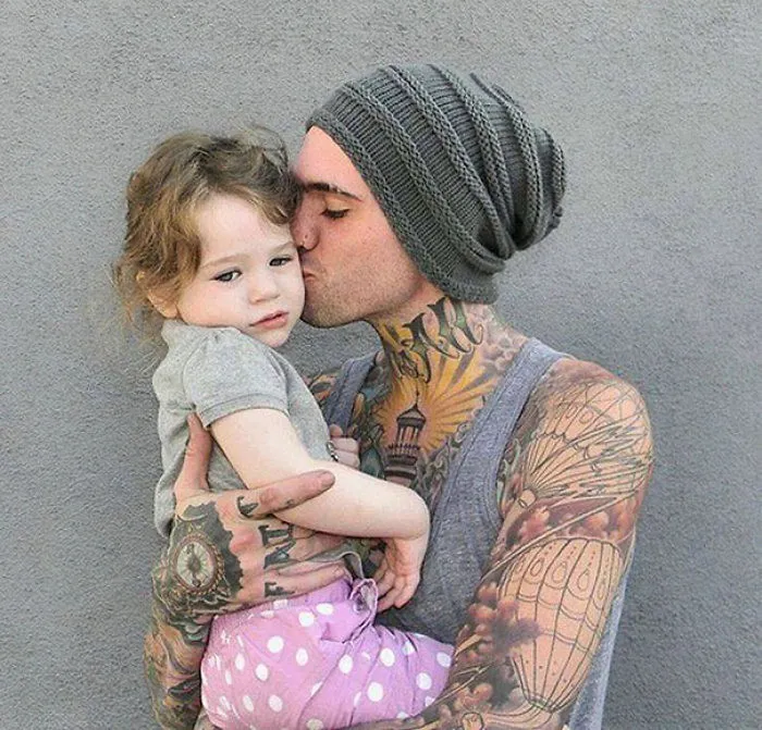 Tattooed parent lovingly holding and kissing a baby, showcasing a beautiful family bond.