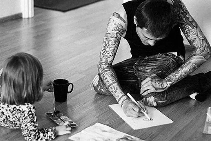 Tattooed parent drawing with child using watercolors on a wooden floor.
