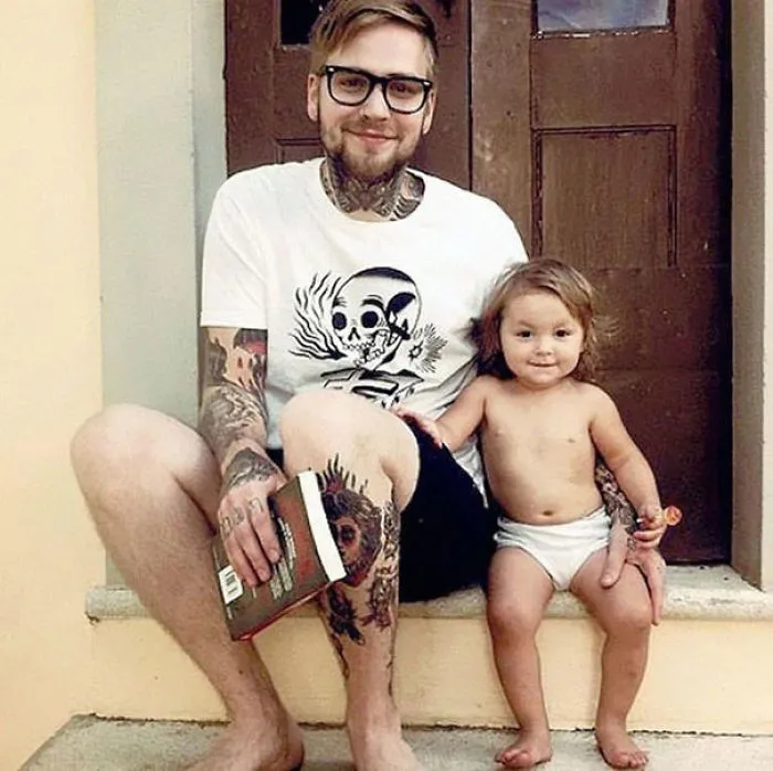 Tattooed parent sitting with child on steps, parent holding book, both smiling at camera.