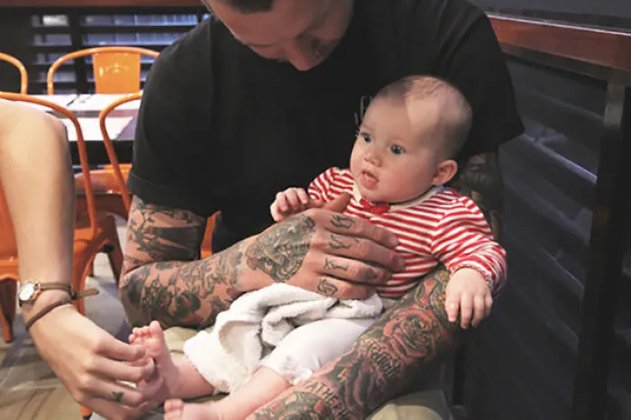 Tattooed parent holding baby, both looking beautiful together in a cozy setting.