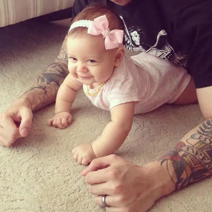 Baby crawling on carpet, next to tattooed parent, both smiling and enjoying time together.