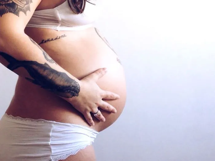 Pregnant tattooed parent gently holding belly, showcasing body art in a serene setting.