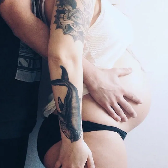 Pregnant person with a tattooed arm embracing belly, showcasing beautiful connection with tattooed parents.