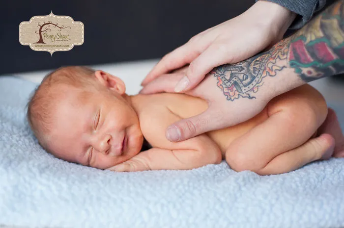 Tattooed parent gently touches their sleeping baby on a blue blanket, showcasing a beautiful family bond.