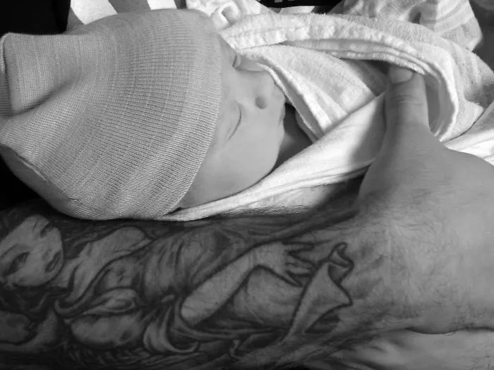 Baby wrapped in a blanket, held by a tattooed parent, showcasing beautiful art and bond together.