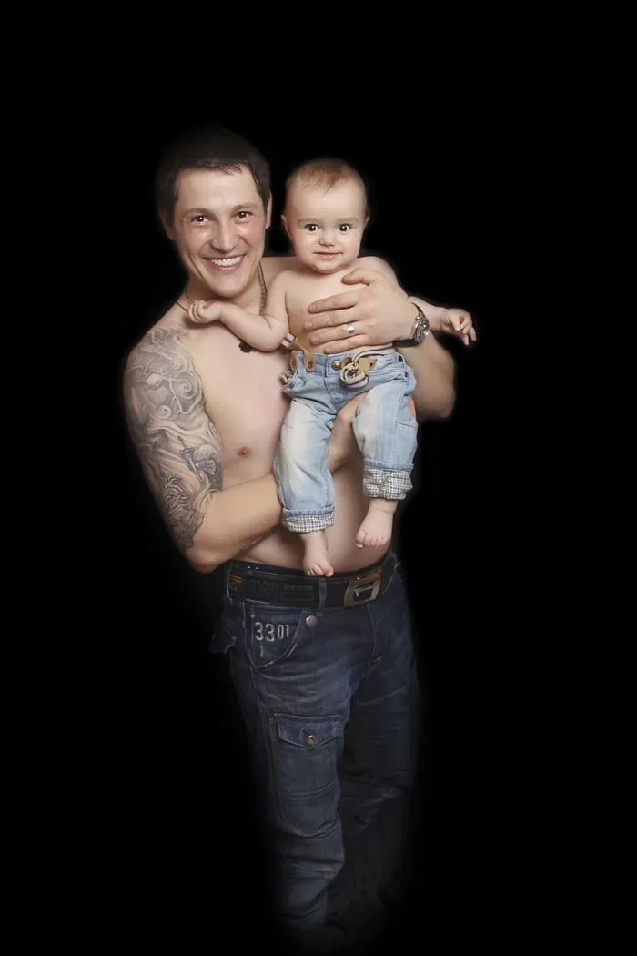 Tattooed parent smiling, holding a baby, both shirtless, on black background.