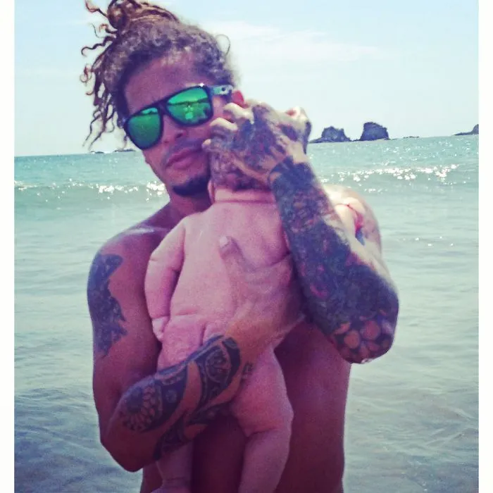 Tattooed parent holding baby at the beach, showcasing beautiful bonding and artistic body art in natural light.