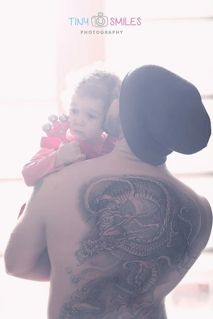 Baby held by tattooed parent with a dragon tattoo, both embracing in soft light.