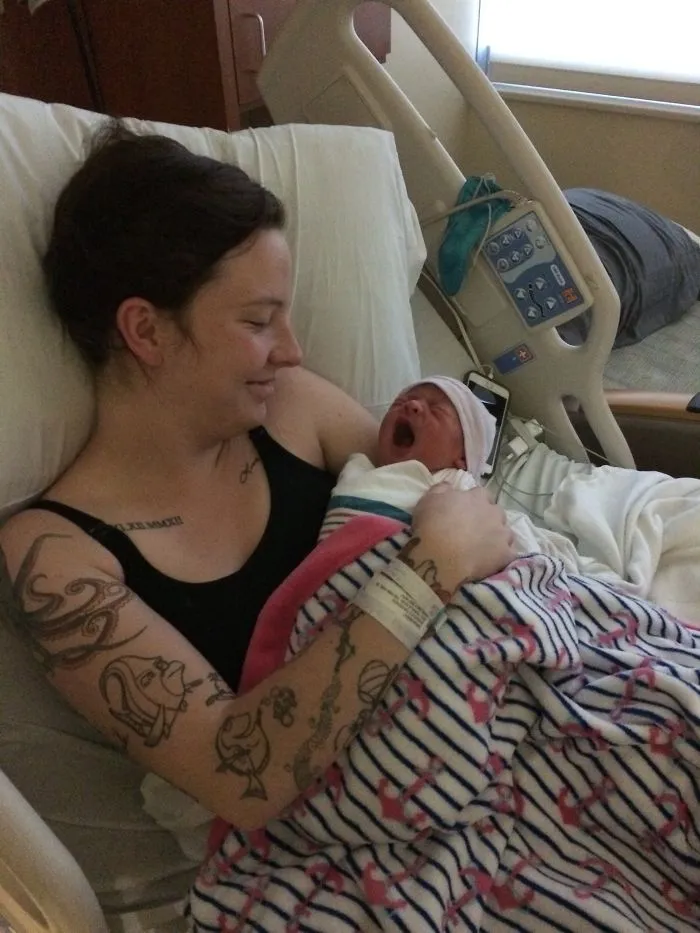 Tattooed parent lovingly holding their newborn baby in a hospital bed, surrounded by soft blankets.