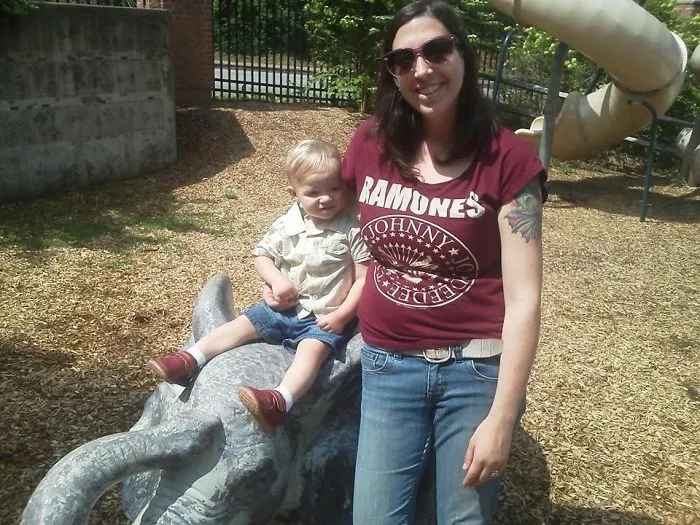 Tattooed parent with child sitting on elephant sculpture at a playground, both smiling in sunny weather.