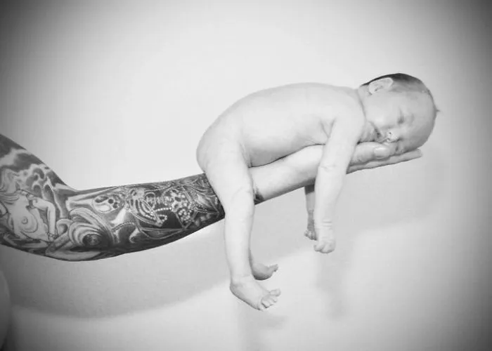 Tattooed parent gently cradles a sleeping baby on their arm, showcasing beautiful ink and a serene moment together.