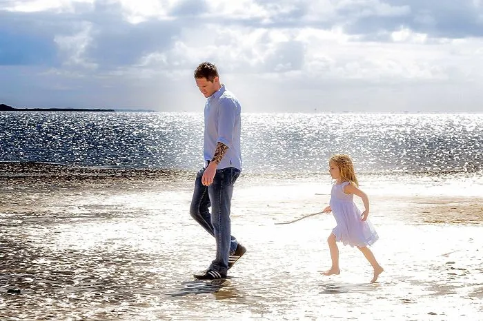 Tattooed parent walking on the beach with their child, enjoying a sunny day together.