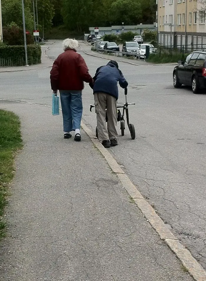 Elderly couple walking on a sidewalk, one using a walker, proving age doesn't limit fun.
