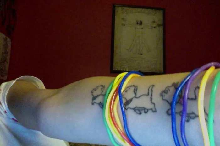 Arm with simple cat tattoos in line art style, adorned with colorful bracelets.