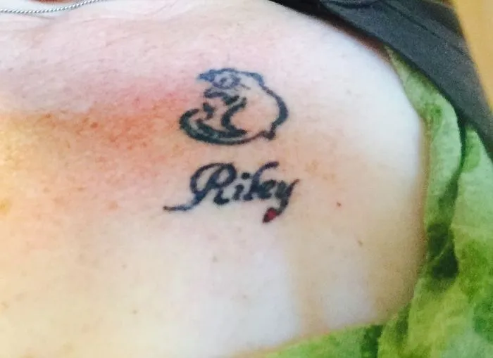 Minimalist cat line art tattoo with name "Riley" beneath it.