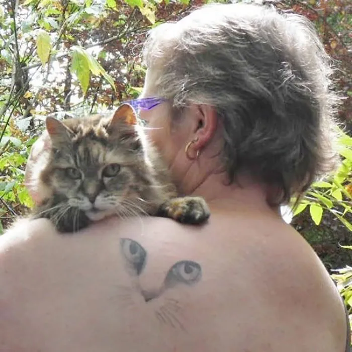 Person holding a cat with a simple cat tattoo on their back amidst greenery.