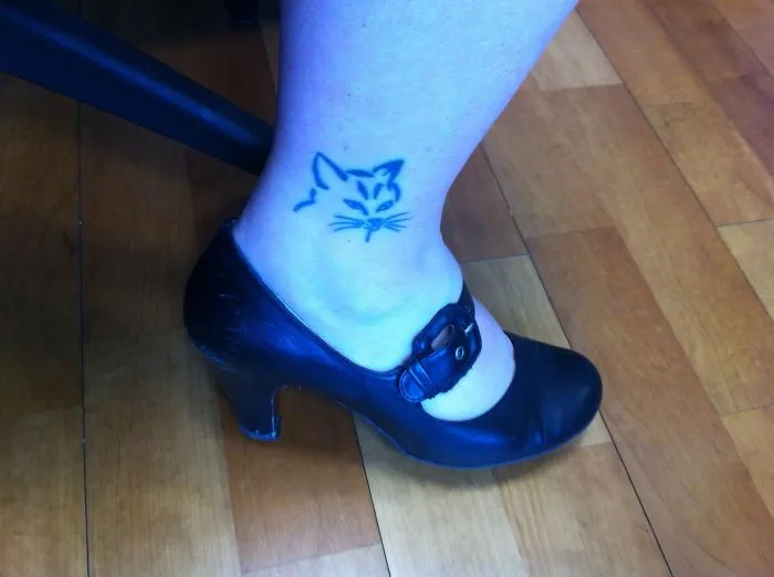 Simple cat tattoo line art on ankle, woman in black heels on wooden floor.