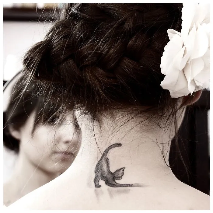 A simple line art cat tattoo on the back of a woman's neck, featuring the cat in a stretching pose.