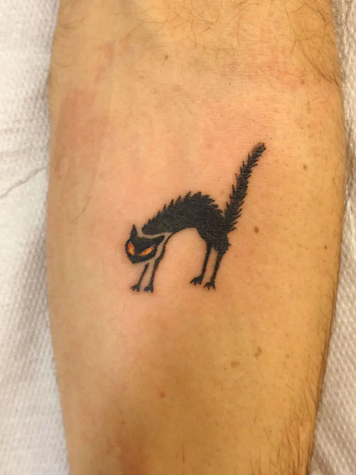 Simple cat tattoo design featuring a stretching black cat with orange eyes on an arm.