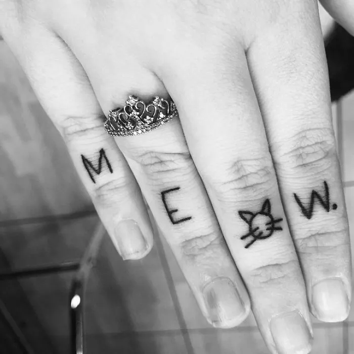 Line art cat tattoo design spelling "MEOW" on fingers with a crown ring.
