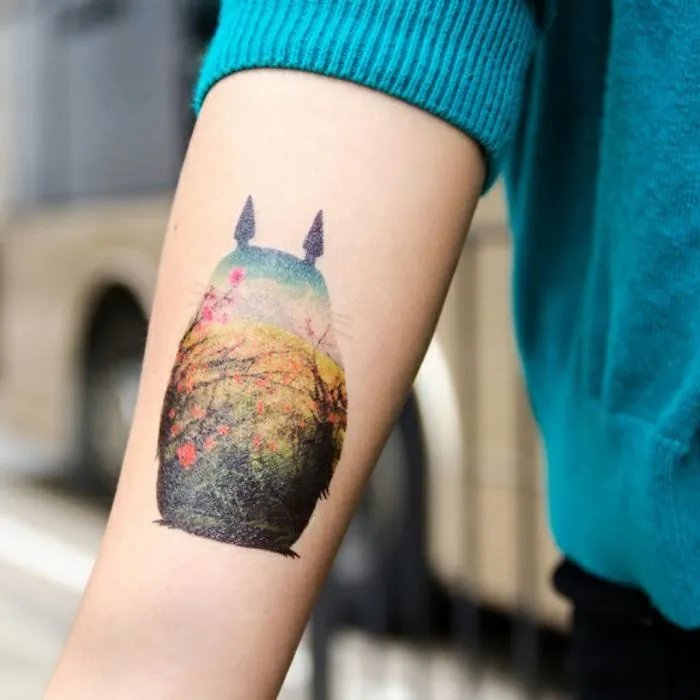 Studio Ghibli Totoro tattoo inspired by Miyazaki films on a person's arm featuring a colorful nature scene.