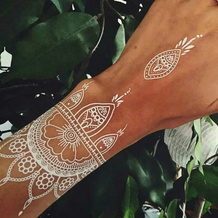 White henna-inspired tattoo resembling elegant lace on a forearm, surrounded by green leaves.