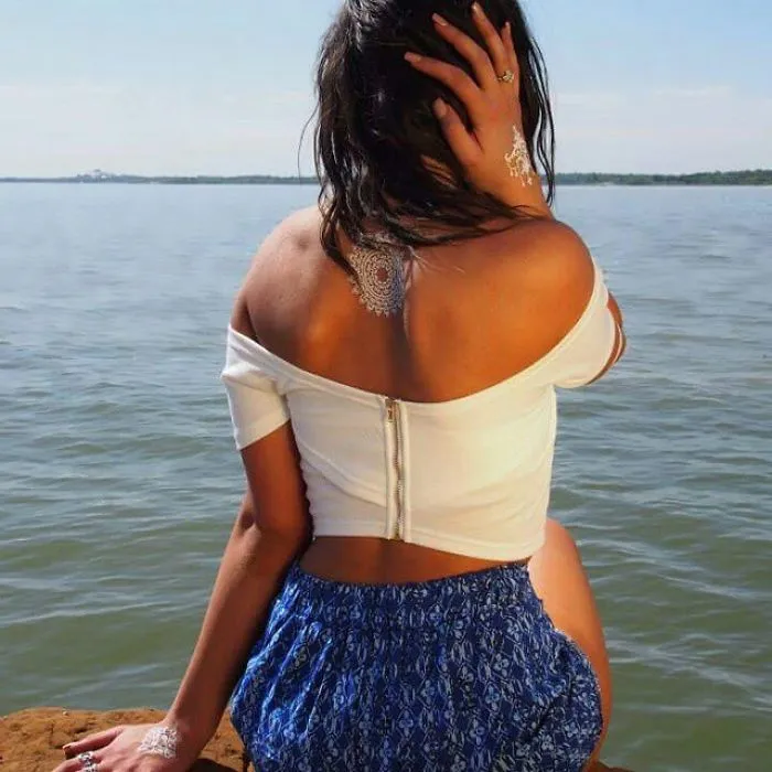Woman with white henna-inspired tattoo on back and hand, wearing off-shoulder top by a lake.