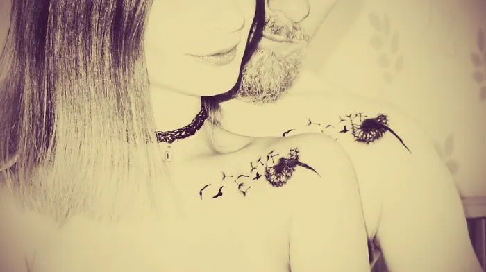 Couple with matching wedding tattoos on their shoulders, featuring dandelions and birds, embracing closely.