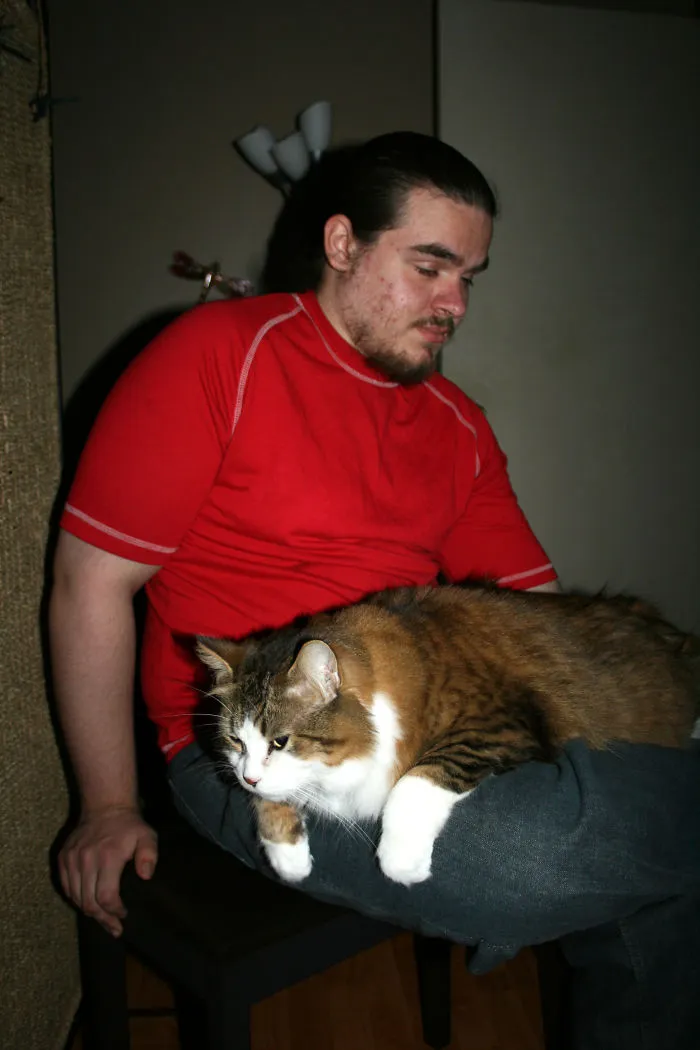 Man in a red shirt sitting with a large Maine Coon cat resting comfortably on his lap indoors.