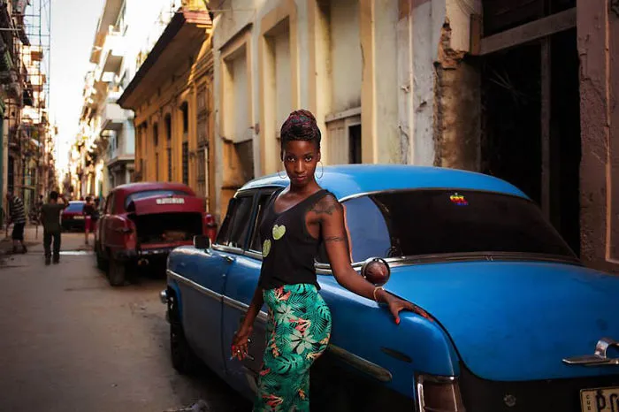 Woman posing beside a blue vintage car on a street, showcasing global beauty by a Romanian photographer.