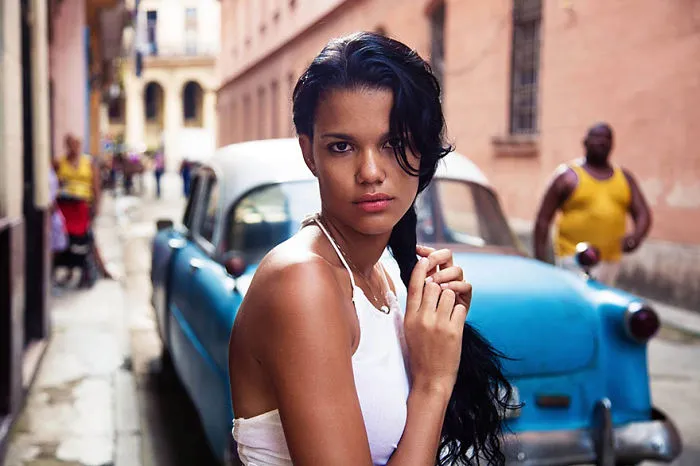 Woman in white top poses in front of a blue vintage car on a city street, photographed by a Romanian photographer.