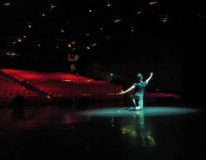 Ballet dancer posing on an empty stage under spotlight, showcasing the art and cost of applause.