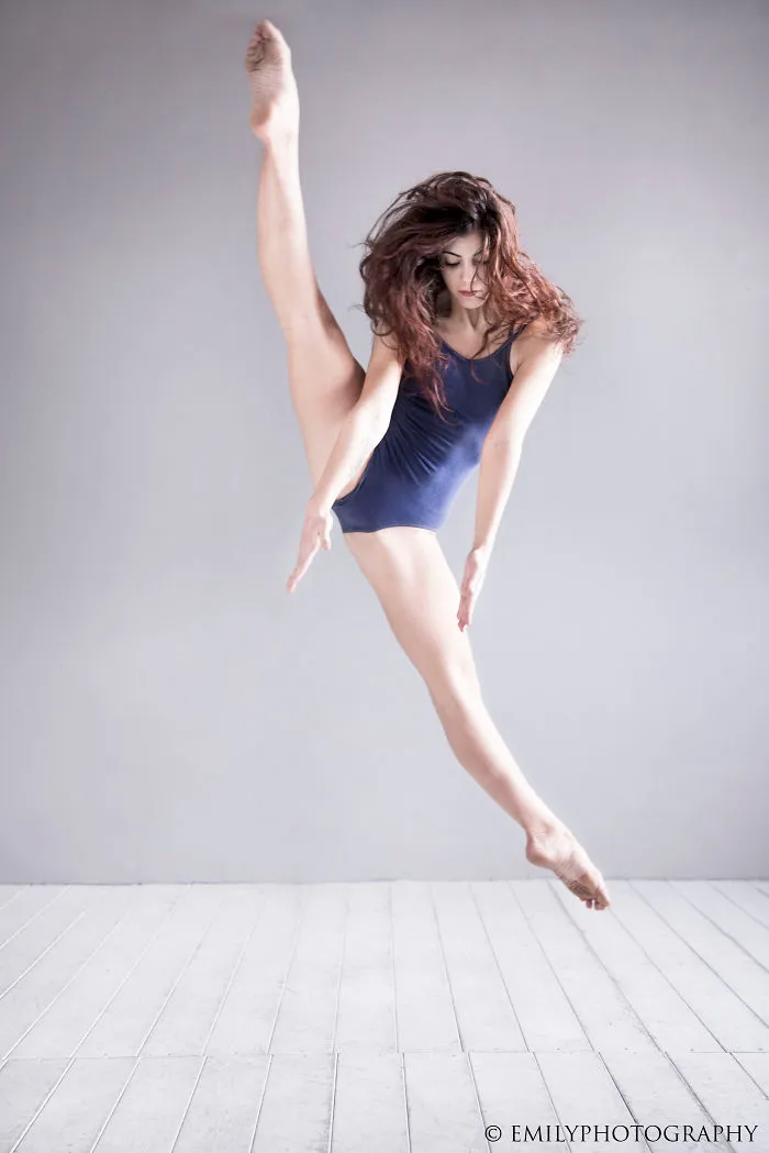 Ballet dancer in mid-air leap on a wooden floor, wearing a navy leotard.