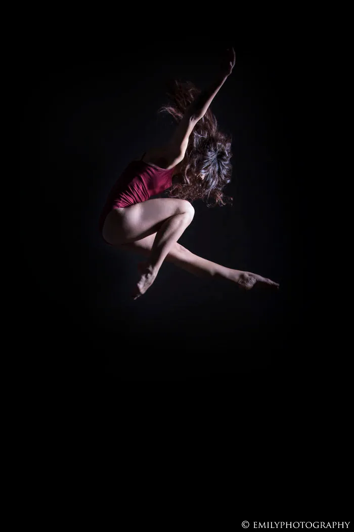 Ballet dancer in a red leotard performing a mid-air leap against a black background, showcasing the art of ballet.