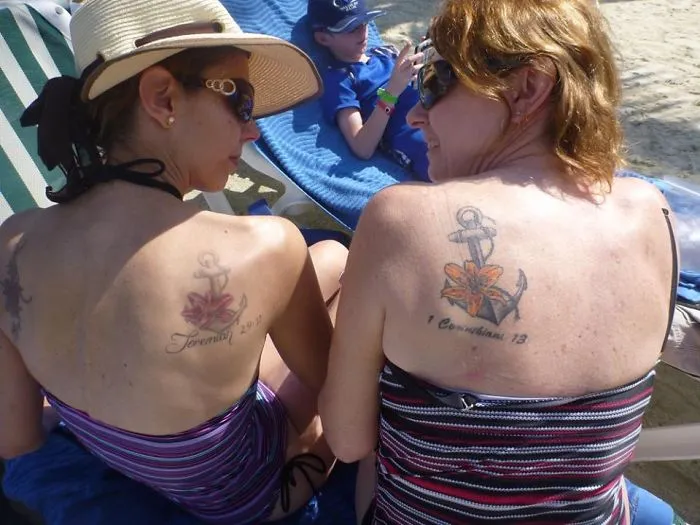 Sisters on the beach showcasing matching anchor and flower tattoos on their backs.