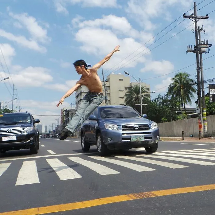 Dancer in mid-air leap on a city street, shirtless with jeans, beside cars, showcasing the art of ballet.