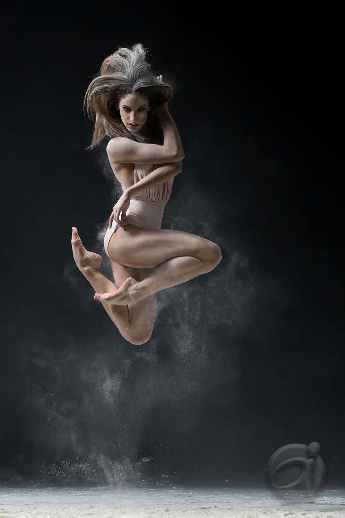 Ballet dancer mid-leap in a dynamic pose, surrounded by floating dust on a dark background.