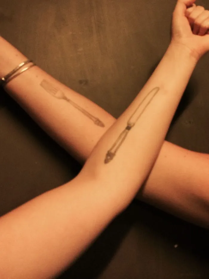 Two forearms crossed with matching spoon and fork tattoos, symbolizing a sister bond.