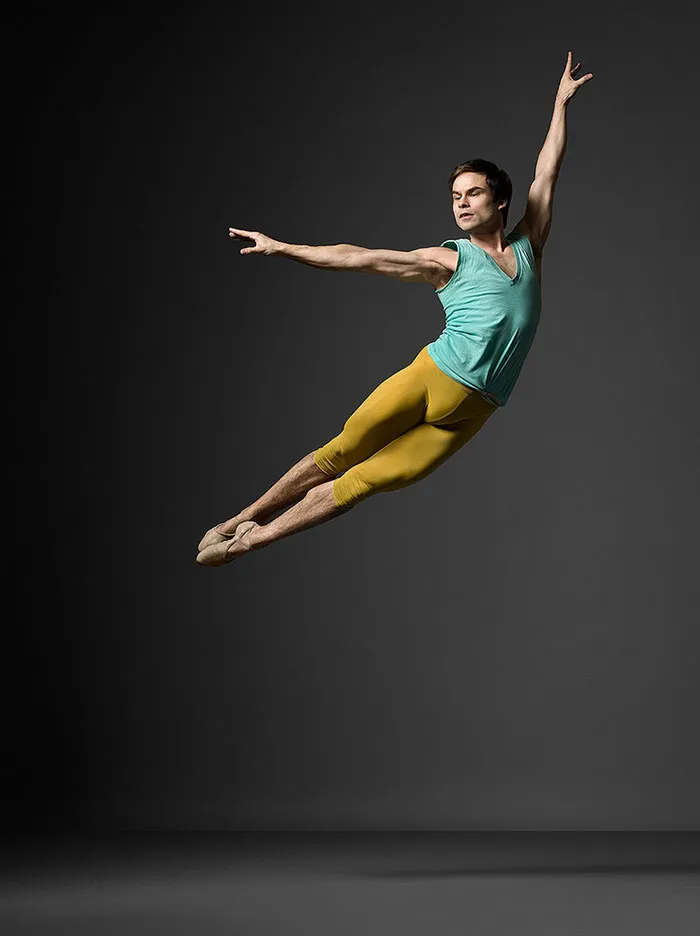 Ballet dancer in mid-air, wearing a teal top and yellow tights, showcasing grace and strength.