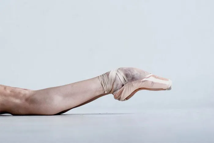 Ballerina's foot in pointe shoe, showcasing the cost of applause in ballet, resting on a smooth white surface.