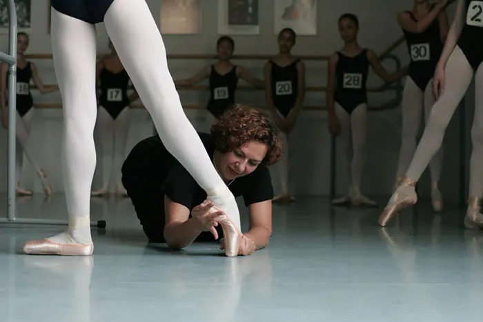 Ballet instructor adjusting a dancer's pointe shoe during rehearsal, focusing on technique and precision.