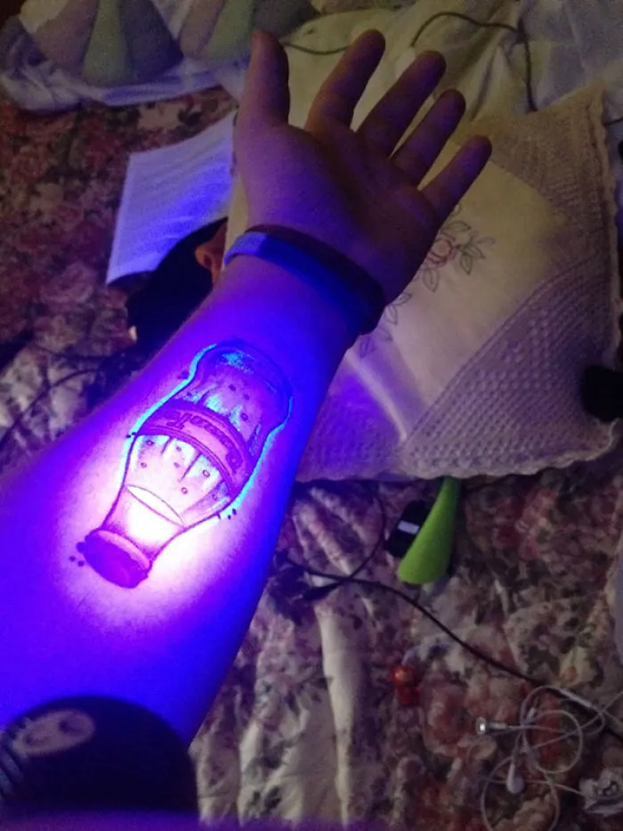 Glow in the dark tattoo of a bottle on forearm, illuminated under black light.
